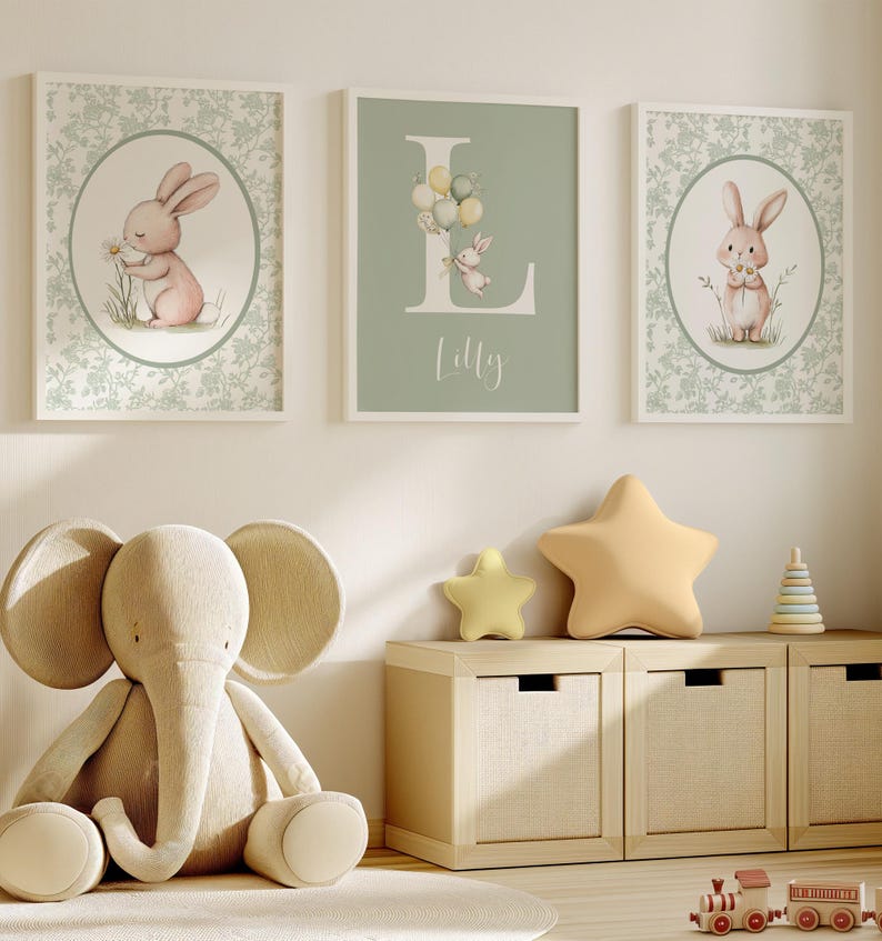 K&ouml;nnte beinhalten: Drei gerahmte Kunstdrucke f&uuml;r das Kinderzimmer mit Hasen und floralen Mustern. Ein Druck zeigt den Buchstaben "L" mit Luftballons und einem Hasen sowie den Namen "Lilly". Ein Pl&uuml;schelefant und sternf&ouml;rmige Kissen befinden sich in der N&auml;he eines Holzschranks.
