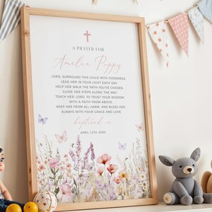Op de afbeelding: Een ingelijste print met een licht houten frame. De print toont een gebed voor "Amelia Poppy" met een bloemenontwerp in zachte roze en paarse tinten. Vlinders en een kruis zijn ook inbegrepen. De woorden "gedoopt op 12 april 20XX" staan onderaan.