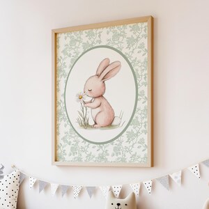 K&ouml;nnte beinhalten: Ein gerahmter Druck mit einer Aquarellillustration eines rosa Kaninchens, das an einer G&auml;nsebl&uuml;mchen schnuppert. Das Kaninchen befindet sich in einem ovalen Rahmen mit hellgr&uuml;nem Rand, vor einem floral gemusterten Hintergrund. Der Rahmen ist hellholzfarben.