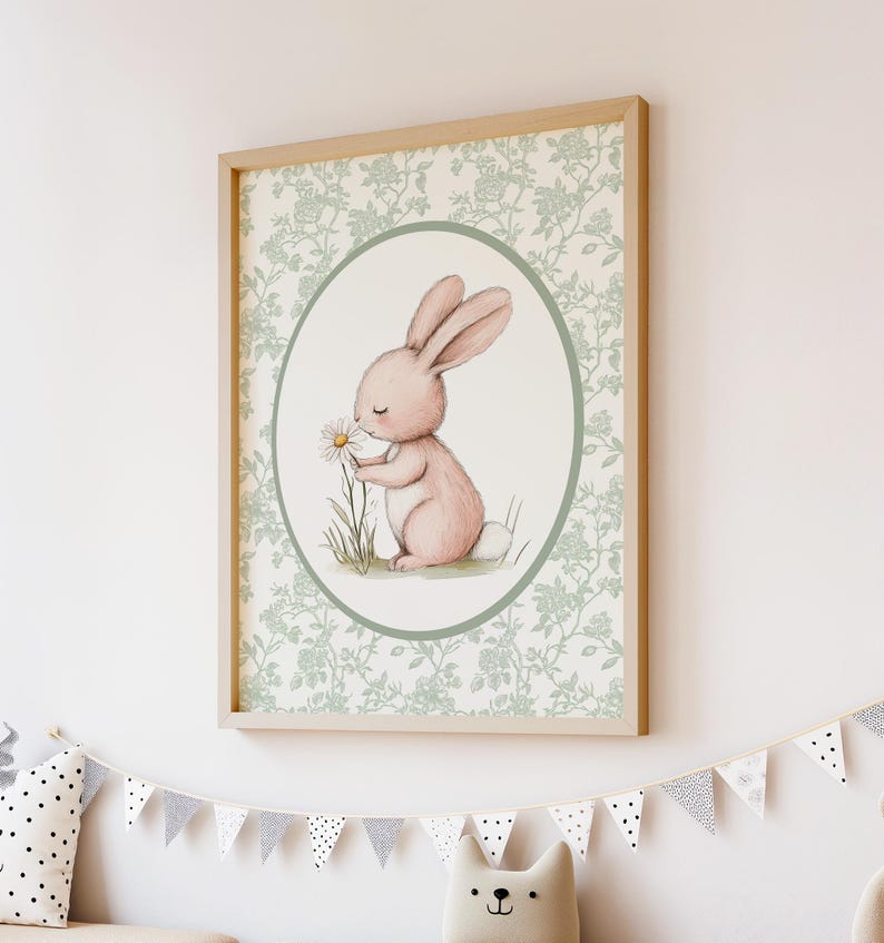 K&ouml;nnte beinhalten: Ein gerahmter Druck mit einer Aquarellillustration eines rosa Kaninchens, das an einer G&auml;nsebl&uuml;mchen schnuppert. Das Kaninchen befindet sich in einem ovalen Rahmen mit hellgr&uuml;nem Rand, vor einem floral gemusterten Hintergrund. Der Rahmen ist hellholzfarben.