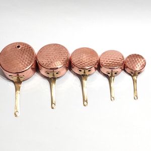 Può includere: Un set di cinque pentole in rame con texture martellata e manici in ottone. Le pentole variano in dimensioni, la più grande ha un diametro di circa 15 cm e la più piccola circa 7,5 cm. Il rame ha una calda tonalità rosso-marrone.