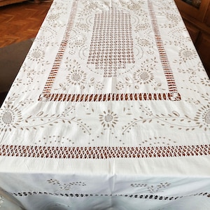 Puede incluir: Mantel rectangular blanco con intrincados diseños florales y geométricos calados. Los bordes y el centro presentan patrones detallados, incluyendo girasoles y filas de pequeños círculos. El mantel está hecho de una tela blanca suave.