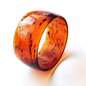 Puede incluir: Una pulsera tipo brazalete ancha y translúcida con un acabado pulido y brillante en un tono ámbar anaranjado cálido. El material muestra un patrón jaspeado con manchas y vetas irregulares de color marrón oscuro distribuidas por toda la forma circular.