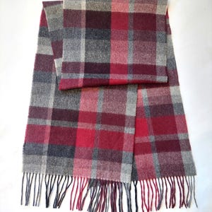 May include: A rectangular scarf with a plaid pattern in shades of red, gray, and burgundy. The scarf has a fringed edge and is made of a soft, woven material. The design features a classic check pattern, suitable for cold weather.