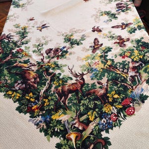 May include: Cream-colored tablecloth featuring a forest scene with deer, birds, and mushrooms. The design includes green foliage, brown deer, and colorful accents. The tablecloth is made of a woven fabric.