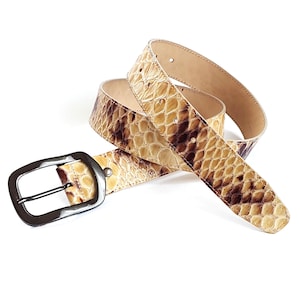 Pode incluir: Um cinto com uma superfície texturizada com padrão de pele de cobra em tons de bege, creme e castanho escuro. O cinto inclui uma fivela retangular de metal escuro e uma série de orifícios perfurados ao longo da tira para ajuste.