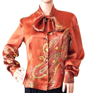 May include: A long-sleeved blouse in a rich copper-orange hue, featuring a pussy-bow neckline and a button-down front. The garment is adorned with a large, intricate paisley pattern in shades of yellow, pink, and green along the front and sleeves.