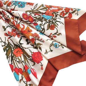 Può includere: Una sciarpa di seta color crema con un vivace motivo floreale. Il design presenta fiori arancioni, rossi e blu con foglie e steli verdi. La sciarpa ha un bordo marrone ed è piegata, mostrando gli intricati dettagli della stampa botanica.