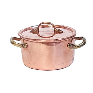 Può includere: Una piccola pentola di rame con coperchio e due manici laterali. La pentola ha una finitura in rame lucido con manici e maniglia del coperchio color ottone. La pentola è progettata per cucinare.