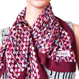 Peut inclure: Un foulard bordeaux orné d'un motif répétitif de lignes verticales reliées par des cercles dans des tons rose, bleu clair et blanc. Le foulard présente une bordure bordeaux unie et une étiquette rectangulaire blanche avec le texte Loredano.