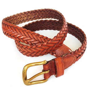 Può includere: Una cintura in pelle intrecciata marrone con una fibbia in ottone lucido. La cintura presenta una trama intrecciata lungo tutto il cinturino e include un passante in pelle vicino alla fibbia, che mostra un piccolo logo in rilievo di un cavaliere.