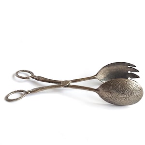 Puede incluir: Pinzas de servir plateadas con mangos ornamentados y extremos para servir. Un extremo tiene forma de cuchara con un diseño grabado, y el otro tiene forma de tenedor. Los mangos tienen lazos ovalados.