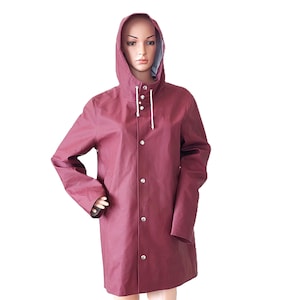 Puede incluir: Una gabardina con capucha de color burdeos con una cremallera y cierre de botones blancos. La gabardina tiene un corte largo y holgado y está hecha de un material impermeable.