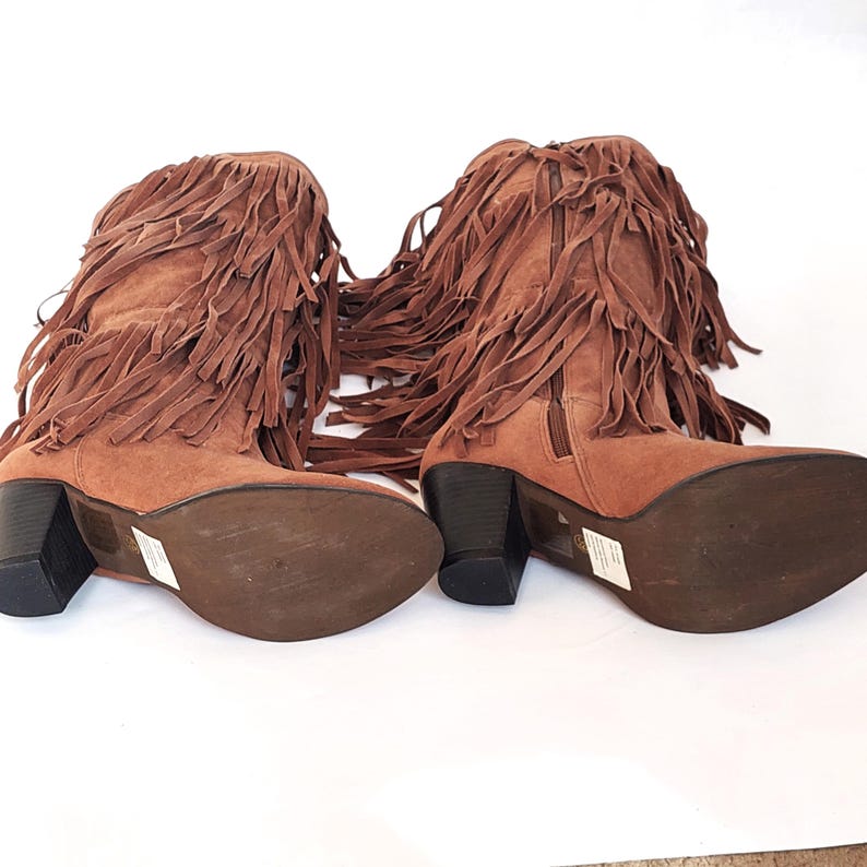 May include: A pair of brown suede boots with a fringe design. The boots have a block heel and a side zip. The fringe detail covers the top of the boots. The boots are set against a white background.