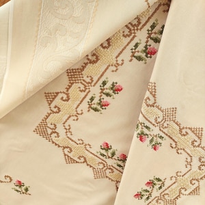 Puede incluir: Mantel de color crema con intrincado bordado de punto de cruz. El diseño presenta un borde de patrones geométricos de color beige y dorado y motivos florales con acentos rojos y verdes. La tela está doblada, revelando la textura y la artesanía detalladas.