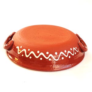May include: A terracotta-colored ceramic bowl with two handles. The rim is decorated with a white wavy pattern. The bowl is round and has a rustic, handmade appearance. The handles are curved and integrated into the bowl's design.