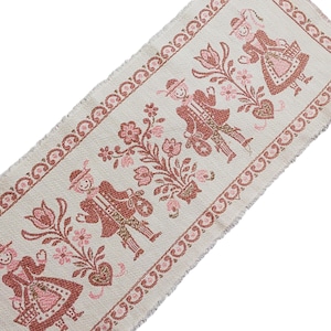 Op de afbeelding: Crèmekleurige stoffen loper met een herhalend patroon van gestileerde figuren in traditionele klederdracht, bloemen en harten in roze en bruine tinten. De randen hebben een decoratieve rand en een gefranjerde afwerking. Het ontwerp roept een volkskunst esthetiek op.