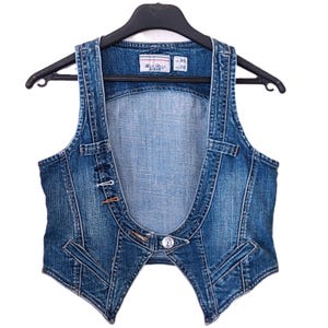 Peut inclure: Un gilet en jean bleu avec un design frontal ouvert unique. Le gilet comporte une fermeture boutonnée, des coutures décoratives et de petites poches. Il est suspendu à un cintre noir. Le gilet a un lavage délavé et une petite étiquette.