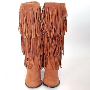 May include: Brown suede knee-high boots with layered fringe detailing. The boots feature a pointed toe and a small heel. The fringe, in shades of brown, drapes down the front, creating a stylish, bohemian look.