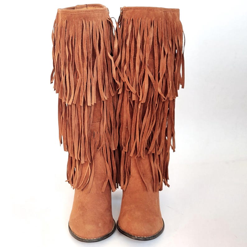 May include: Brown suede knee-high boots with layered fringe detailing. The boots feature a pointed toe and a small heel. The fringe, in shades of brown, drapes down the front, creating a stylish, bohemian look.