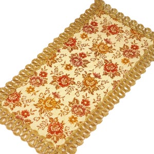 Può includere: Runner da tavolo rettangolare floreale con sfondo crema e un motivo di rose rosse e arancioni. I bordi sono rifiniti con una frangia dorata ad anello. Il runner è probabilmente realizzato in tessuto.