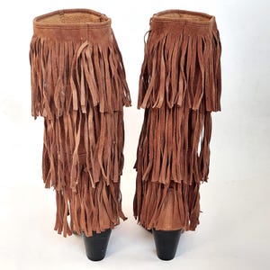 May include: A pair of brown suede boots with multiple layers of long fringe. The boots have a short heel and a soft, textured appearance. The fringe cascades down the front and sides of the boots.