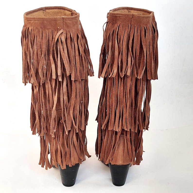 May include: A pair of brown suede boots with multiple layers of long fringe. The boots have a short heel and a soft, textured appearance. The fringe cascades down the front and sides of the boots.
