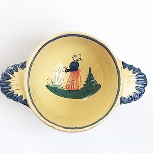 May include: A yellow ceramic bowl with blue and white trim and two handles. The bowl features a hand-painted scene of a woman in a blue dress and red skirt standing in a field with a tree and flowers.