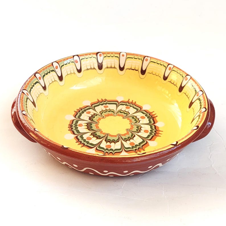 May include: A round, ceramic bowl with a yellow interior and a brown exterior. The bowl features a decorative floral pattern in the center and along the rim, with green, orange, and white accents. It has two small handles on either side.