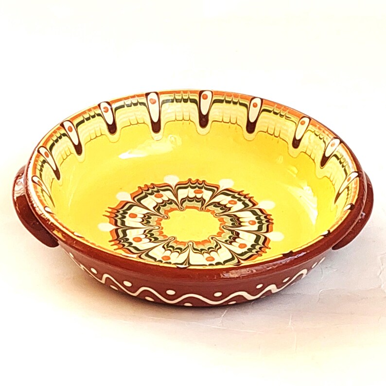 May include: A round, ceramic bowl with a yellow interior and a dark red exterior. The bowl features a floral design in the center and decorative patterns around the rim. Two small handles are on either side of the bowl.