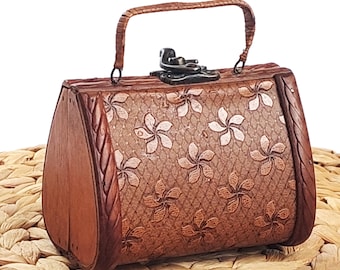 May include: A small brown wooden handbag featuring a textured surface with a repeating floral pattern. The bag has a curved handle, a dark metal latch closure on top, and smooth wooden side panels with a braided edge detail.