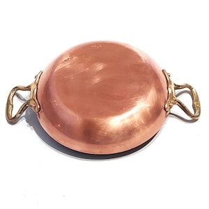 May include: A copper-colored, round cooking pot with two brass handles. The pot has a shiny, reflective surface and is set against a white background. The handles are curved and attached to the sides of the pot.