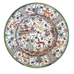 May include: A round ceramic plate featuring a hand-painted design with a central medallion and a wide border. The artwork depicts various animals including deer and birds, interspersed with floral motifs, vines, and geometric grid patterns.