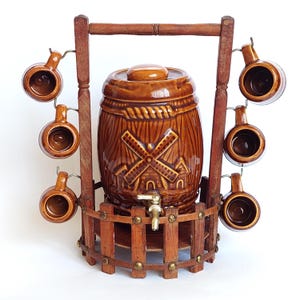 Op de afbeelding: Een bruine keramische vaten-drankdispenser met een windmolenreliëf en een metalen kraan, geplaatst in een houten standaard met een hekachtige basis en zes bijpassende bruine keramische mokken die aan haken aan de zijsteunen hangen.