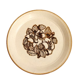 Può includere: Un piatto rotondo in ceramica bianco sporco con un motivo floreale marrone con fragole e fiori. Il piatto ha una consistenza macchiettata e un bordo marrone. Il motivo floreale è al centro.