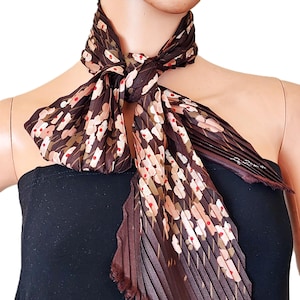 May include: A dark brown pleated scarf featuring a repeating floral pattern in shades of light pink, white, and small red dots. The scarf is tied in a knot around the neck and has a textured, accordion-pleated finish with frayed edges.