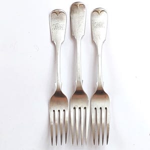 May include: Three antique silver forks with heart-shaped tops and engraved initials. The forks have tines and long, slender handles. The initials are different on each fork. The forks are arranged vertically against a white background.