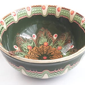 Op de afbeelding: Een decoratieve keramische kom met een donkergroen interieur en exterieur. De rand heeft een herhalend patroon van groene, roze en witte golvende lijnen. De binnenkant van de kom heeft een bloemmotief in tinten oranje, groen en rood.