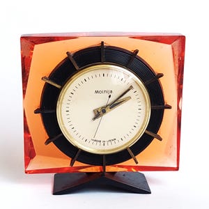 May include: A square-shaped Molnija desk clock encased in translucent orange resin. The clock features a circular black frame with radiating spokes, a cream-colored face with black markers, gold-toned hands, and the text Molnija and Made in USSR.