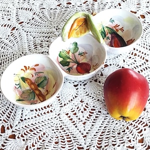 Puede incluir: Una fuente de cerámica blanca con tres cuencos conectados, cada uno con un diseño de acuarela de verduras. Una manzana roja se encuentra junto a la fuente sobre un mantel de encaje blanco. La fuente tiene un asa con un diseño de hoja.