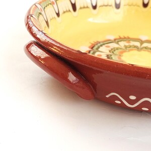May include: A close-up of a ceramic bowl with a yellow interior and a reddish-brown exterior. The bowl has a decorative floral pattern around the rim and a handle on the side. The bowl is on a white surface.