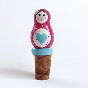 May include: A pink and white nesting doll bottle stopper with a blue heart design. The stopper is made of wood and has a cork base.