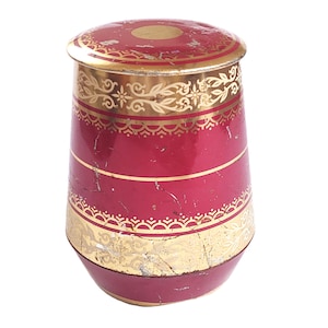 Könnte beinhalten: Eine zylindrische Vintage-Blechdose mit Deckel. Die Dose ist überwiegend tiefrot, akzentuiert mit goldenen Bändern und dekorativen Mustern. Der Deckel hat einen goldenen Kreis in der Mitte. Die Dose weist Gebrauchsspuren auf, was ihren antiken Charme unterstreicht.