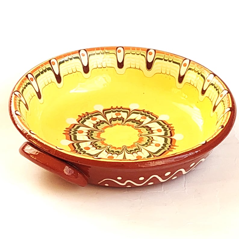 May include: A decorative ceramic bowl with a vibrant yellow interior and a detailed floral pattern in the center. The bowl has a deep red exterior with white and brown accents, and a small handle. The rim features a repeating design.