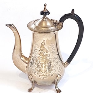 Puede incluir: Cafetera antigua de color plateado con detalles florales y de volutas ornamentados. Cuenta con un asa negra y un adorno decorativo en la tapa. La cafetera tiene tres patas pequeñas y un pico curvo.