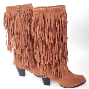May include: A pair of brown suede boots with multiple layers of fringe. The boots have a zipper closure on the side and a small heel. The boots are approximately 30 cm tall.
