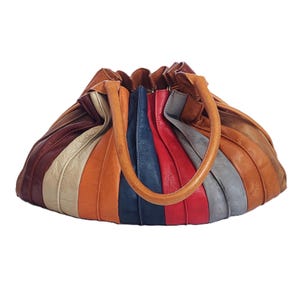 Può includere: Borsa a mano colorata in stile vintage dalla forma arrotondata. La borsa presenta pannelli verticali in vari colori, tra cui marrone, beige, arancione, blu, rosso e grigio. Ha un manico marrone chiaro.