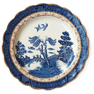 May include: A blue and white decorative plate with a scalloped edge and gold trim. The plate features a detailed landscape scene with a bridge, trees, and buildings. The outer border has a geometric pattern.