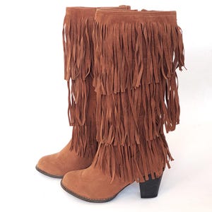 May include: A pair of brown suede knee-high boots with multiple layers of long fringe. The boots have a small heel and a rounded toe. The boots are set against a white background.