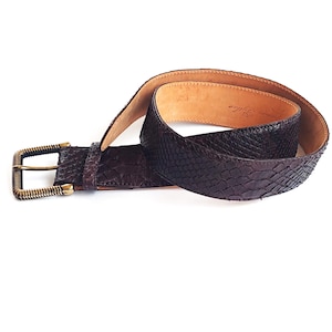 以下が含まれることがあります： 蛇柄のブラウンレザーベルトと真鍮製のバックル。ベルトは半分に折り畳まれ、バックルは画像の左側にある。
